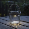 Szklany lampion solarny LED z jutowym uchwytem ustawiony na drewnianym stole tarasowym po zmroku, emitujący ciepłe białe światło.