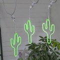 Zbliżenie na trzy neonowe dekoracje LED w kształcie kaktusa, emitujące intensywne zielone światło. Elementy zamontowane w białych oprawkach E27, widoczne przewody girlandy i roślina w tle.