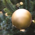 Zbliżenie na matową, złotą bombkę choinkową zawieszoną na gałązce sztucznego igliwia. W tle widoczne są delikatne światełka LED i miękkie, świąteczne rozmycie.