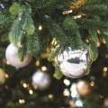 Zbliżenie na choinkowe gałązki z zawieszonymi srebrnymi bombkami – matową i błyszczącą lustrzaną, w której odbija się wnętrze pomieszczenia. Między gałązkami widoczne ciepłe światełka LED.