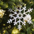 Zbliżenie na dużą, brokatową śnieżynkę w srebrnym kolorze zawieszoną na choince wśród zielonych gałązek, złotych i srebrnych bombek oraz ciepłych lampek LED.