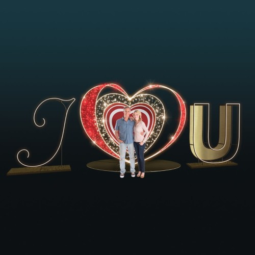 Fotopunkt ‘I ❤ U’ z podświetlanym sercem LED na środku, po bokach litery I i U, ciepłe białe i czerwone światło LED, para stojąca wewnątrz serca, dekoracja na ciemnym tle.