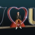 Czerwone serce 3D z jasnym obrysem LED, stanowiące centralny element napisu „I LOVE YOU”; mężczyzna siedzący na podstawie serca, widoczna metalowa konstrukcja i świetlne detale.