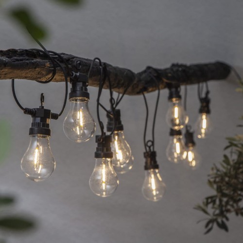 Girlanda ogrodowa LED z przezroczystymi żarówkami w stylu filament zawieszona na gałęzi drzewa, emitująca ciepłe światło w ogrodowej aranżacji.