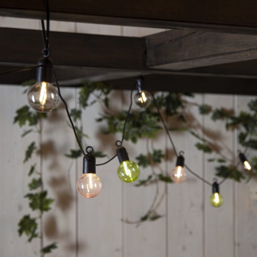 Girlanda ogrodowa LED FIESTA z pastelowymi kloszami zawieszona pod drewnianą pergolą, kolorowe żarówki LED emitujące ciepłe światło, dekoracyjne oświetlenie tarasu w otoczeniu zieleni.