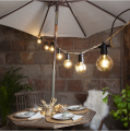 Girlanda żarówkowa E27 SYSTEM 24 z ciepłymi żarówkami LED 24V zawieszona nad stołem ogrodowym pod parasolem. Nastrojowe oświetlenie tarasu tworzące przytulną atmosferę do kolacji na świeżym powietrzu.