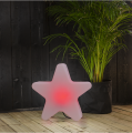Dekoracyjna lampa w kształcie gwiazdy świecąca czerwonym światłem ustawiona na drewnianym tarasie obok rośliny doniczkowej.