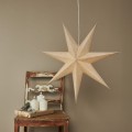 Beżowa gwiazda świąteczna o przestrzennej, geometrycznej formie, zawieszona nad drewnianym krzesłem ze świecami, w przytulnej aranżacji wnętrza w stylu vintage.