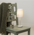 Lampa stołowa z abażurem ustawiona na zielonym krześle obok szklanego wazonu z gałązkami i koca, przytulna aranżacja wnętrza z ciepłym światłem żarówki LED E14.
