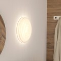 Okrągła lampa sufitowa BATTISTONA z pilotem, zamontowana na jasnej ścianie i emitująca ciepłe światło wokół białej oprawy.