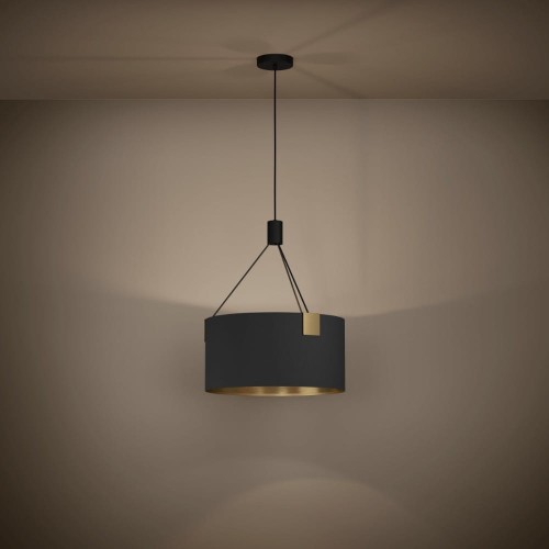 Lampa wisząca TORTOLA 1 z czarnym cylindrycznym kloszem z tkaniny i mosiężnymi detalami, zawieszona na tle ciemnej ściany i emitująca ciepłe światło.