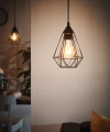 Na tle jasnej ściany lampa wisząca TARBES prezentuje smukły ażurowy klosz z czarnych metalowych prętów, otaczający żarówkę E27 emitującą ciepłe światło.