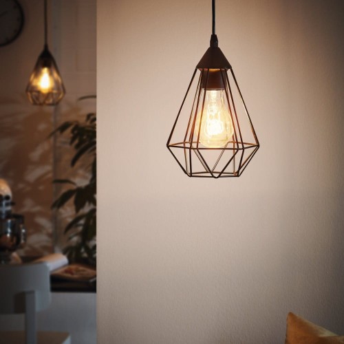 Lampa wisząca TARBES z czarnym metalowym kloszem o ażurowej, geometrycznej konstrukcji, zawieszona na tle jasnej ściany z widoczną świecącą żarówką.