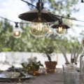 Girlanda ogrodowa z dekoracyjnymi żarówkami LED G45 zawieszonymi pod metalowymi daszkami nad stołem na tarasie, ciepłe światło w ogrodowej aranżacji.