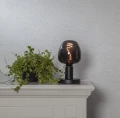 Dekoracyjna żarówka LED filament z przydymionego szkła ustawiona na czarnej lampie stołowej obok rośliny doniczkowej. Ozdobne oświetlenie emitujące ciepłe światło dekoracyjne.