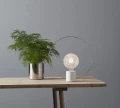 Nowoczesna lampa stołowa z kulistą żarówką LED E27 o strukturalnym kloszu, ustawiona na drewnianym blacie obok rośliny, minimalistyczna aranżacja wnętrza.