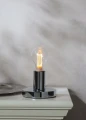 Lampa stołowa z chromowaną podstawą i żarówką LED filament E27, ustawiona na białym meblu.