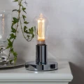 Dekoracyjna żarówka LED filament ST64 w lampie stołowej z chromowaną podstawą, emitująca ciepłe światło w aranżacji wnętrza.
