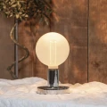 Lampa stołowa z kulistą żarówką LED filament G125 emitującą ciepłe białe światło ustawiona na tkaninie na tle drewnianej ściany.
