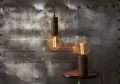 Wisząca lampa z dekoracyjną żarówką LED w złotej klatce oraz lampa stołowa z taką samą żarówką na tle industrialnej ściany metalowej.