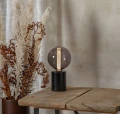 Lampa stołowa z dekoracyjną kulistą żarówką LED filament w przydymionej bańce ustawiona na drewnianym stole obok suszonych roślin.