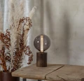 Lampa stołowa z dekoracyjną kulistą żarówką LED filament w przydymionej bańce ustawiona na drewnianym postumencie na drewnianym stole.