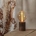 Lampa stołowa z dekoracyjną kulistą żarówką LED filament w bursztynowej bańce ustawiona na drewnianym stole obok suszonych roślin.