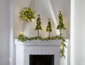 Stylowa dekoracja kominka z trzema smukłymi choinkami LED i zieloną girlandą ze światełkami, zawieszoną poziomo i pionowo przy kominku.