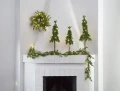 Dekoracja świąteczna z trzema choinkami LED ustawionymi na kominku i zieloną girlandą oświetloną lampkami, zawieszoną nad i wokół kominka.