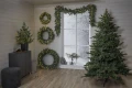 Choinka GREYLAND ustawiona w białym, zimowym wnętrzu z dekoracjami ściennymi z igliwia i girlandą LED wokół okna.