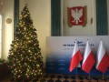Oświetlona choinka z czerwono-złotymi bombkami w holu z godłem Polski, trzema flagami biało-czerwonymi i tablicą informacyjną PKP Polskie Linie Kolejowe S.A.