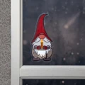 Świąteczna naklejka LED przedstawiająca skrzata w czerwonej czapce trzymającego zapaloną świecę, umieszczona na szybie okna z zimowym, ciemnym tłem.