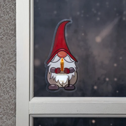 Świąteczna naklejka LED przedstawiająca skrzata w czerwonej czapce trzymającego zapaloną świecę, umieszczona na szybie okna z zimowym, ciemnym tłem.