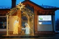 Szopka świetlna 3D z postaciami Świętej Rodziny, ustawiona przed Gminnym Ośrodkiem Kultury i Biblioteką w Polańczyku, oświetlona dekoracjami świątecznymi na tle budynku z zimowym krajobrazem.