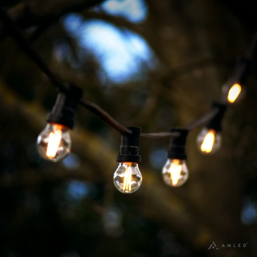 Girlanda ogrodowa z małymi żarówkami LED z serii Soft Glow G45 1,5W 2100K, zawieszona na czarnym przewodzie, widoczna na tle rozmytego krajobrazu z drzewami – idealna do dekoracji przestrzeni zewnętrznej.