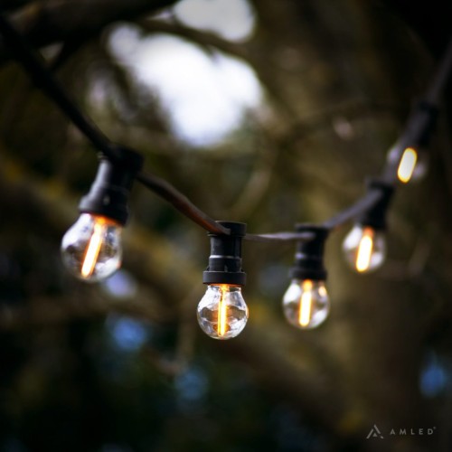 Girlanda ogrodowa z małymi żarówkami LED z serii Soft Glow G45 0,8W 2100K, zawieszona na czarnym przewodzie, widoczna na tle rozmytego krajobrazu z drzewami – idealna do dekoracji przestrzeni zewnętrznej.