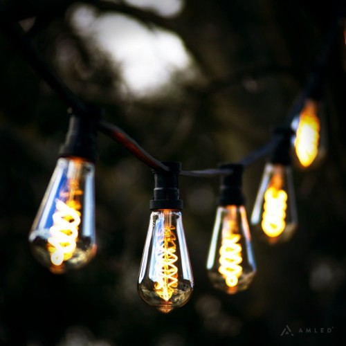Girlanda ogrodowa z żarówkami ST64 3,2W 2100K z serii Decoled Amber w bursztynowym szkle, zawieszona na czarnym przewodzie wśród gałęzi. Żarówki emitują ciepłe światło i mają spiralny żarnik LED.