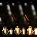 Girlanda ogrodowa z żarówkami LED A60 6,5W 2100K z 3-stopniowym ściemnianiem, zawieszona na czarnym kablu wśród drzew. Żarówki w przezroczystych bańkach świecą ciepłym, nastrojowym światłem.