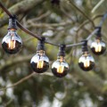 Girlanda ogrodowa z dekoracyjnymi żarówkami LED z serii Plain Smoke G80 o mocy 1,8W, wykonanymi z przydymionego szkła. Żarówki emitują ciepłe, nastrojowe światło i zawieszone są na czarnym przewodzie z czarnymi oprawkami wśród drzew w plenerowej scenerii.