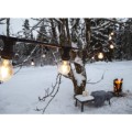Girlanda ogrodowa LED z żarówkami A55 2,4W świeci na tle ośnieżonego ogrodu. Ciepłe światło żarówek kontrastuje z zimowym krajobrazem, w tle widać drzewo, ławkę z kocem i świecami oraz palenisko z ogniem.
