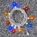 Girlanda ogrodowa z kolorowymi żarówkami LED A60 1W na białym kablu, zwinięta w krąg i ułożona na szarym żwirze. Żarówki świecą w kolorach: niebieskim, czerwonym, żółtym i pomarańczowym, tworząc dekoracyjne oświetlenie.