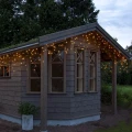 Kurtyna Sople LED zamontowana na drewnianym domku ogrodowym, ciepłe światło 2700 K tworzy nastrojową iluminację na zewnątrz.