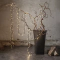 Dekoracyjne girlandy świetlne zwisające z gałęzi ozdobnego, suchego drzewa w dużej donicy, ustawionej w surowym wnętrzu z betonową ścianą.