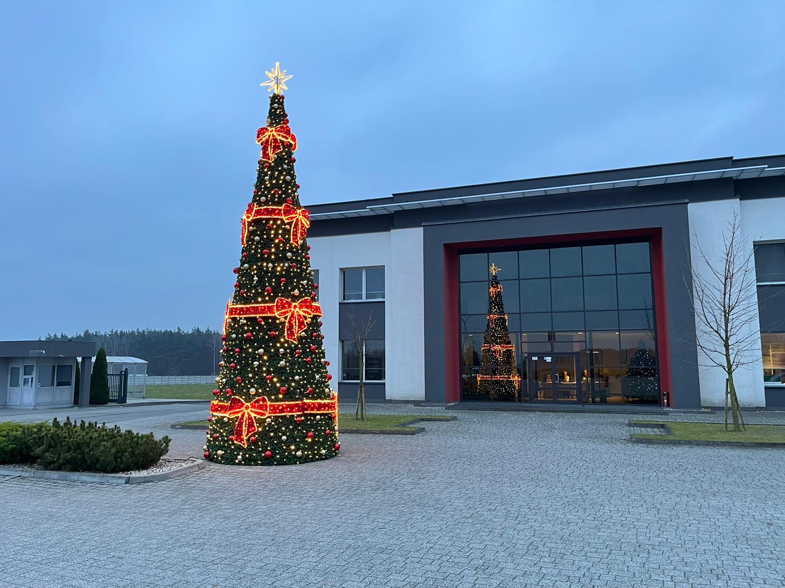 Wysoka stożkowa choinka LED z czerwonymi kokardami i bombkami, ustawiona przed nowoczesnym budynkiem biurowym o świcie. Drzewko udekorowane światłami i ozdobami tworzy efektowną świąteczną aranżację na zewnątrz.