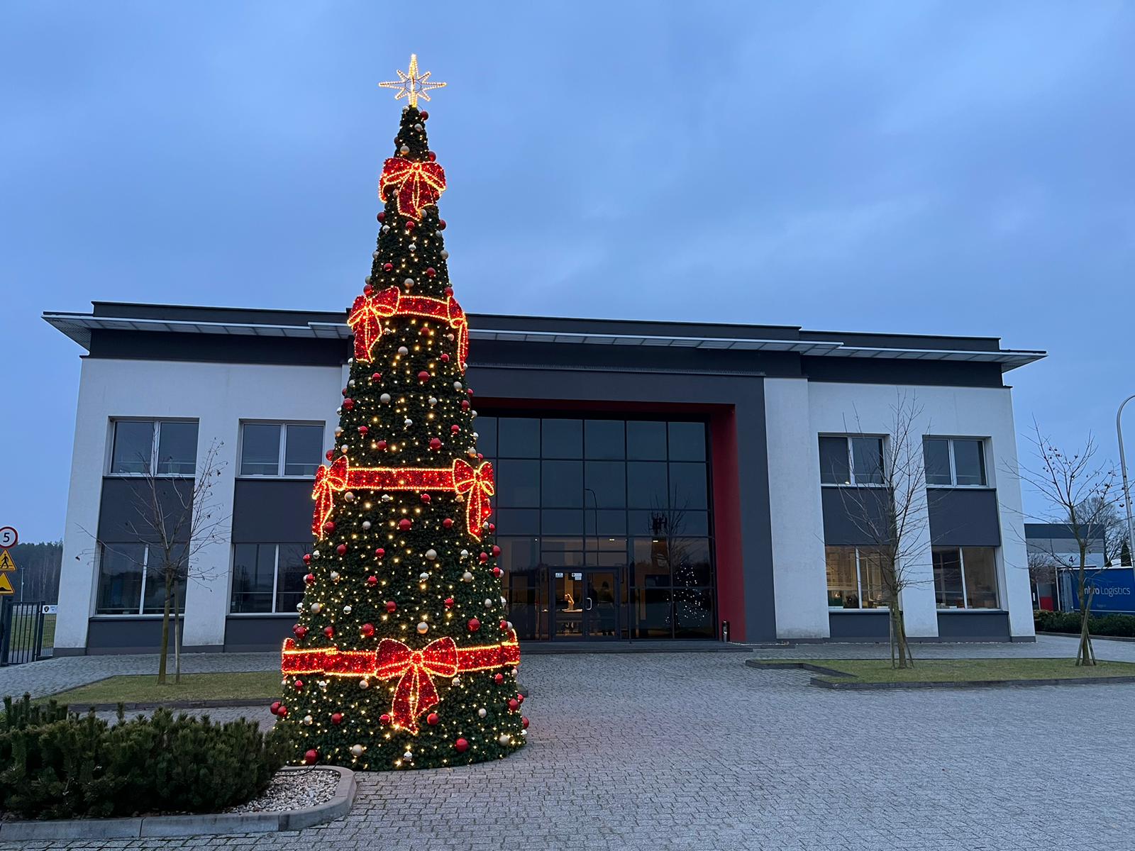 Wysoka stożkowa choinka LED z czerwonymi kokardami i bombkami, ustawiona przed nowoczesnym budynkiem biurowym o świcie. Drzewko udekorowane światłami i ozdobami tworzy efektowną świąteczną aranżację na zewnątrz.
