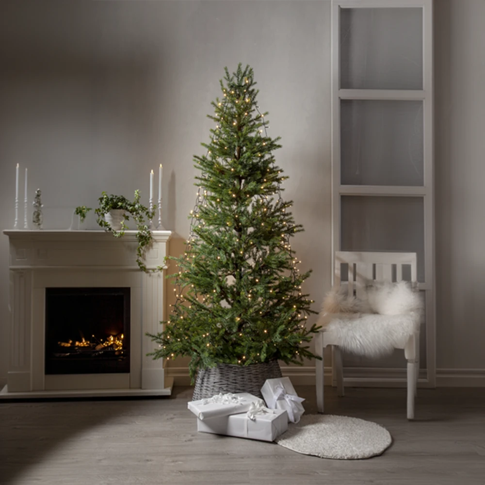 Choinka wewnętrzna z gęstym, śnieżonym igliwiem, ozdobiona ciepłymi światełkami LED, z prezentami i koszem ułożonymi na okrągłym dywaniku.