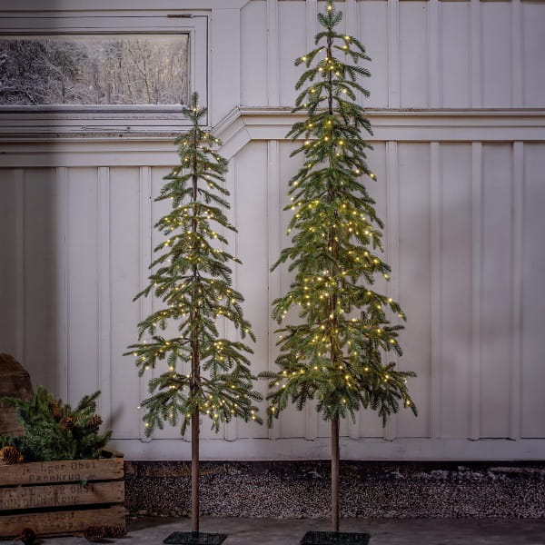 Smukła, minimalistyczna choinka sztuczna typu skandynawskiego, oświetlona ciepłym światłem LED, ustawiona na kwadratowej podstawie, we wnętrzu w stylu rustykalnym.