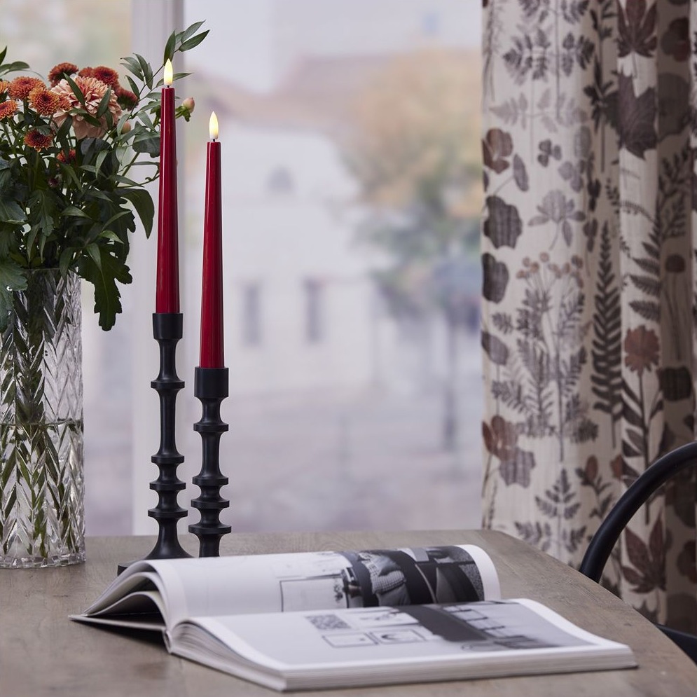 Czerwone smukłe świece LED w szklanych świecznikach obok wazonu z kwiatami na stole przy oknie.
