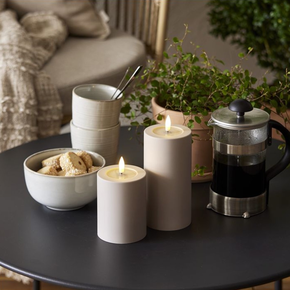 Beżowe świece LED walcowe obok filiżanek, ciastek i kawiarki na stoliku w otoczeniu zielonych roślin.