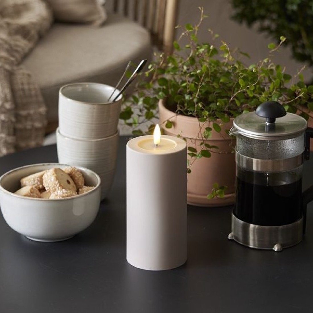 Beżowa świeca LED na stole obok filiżanek, ciasteczek, kawiarki i doniczkowej rośliny w przytulnym wnętrzu.